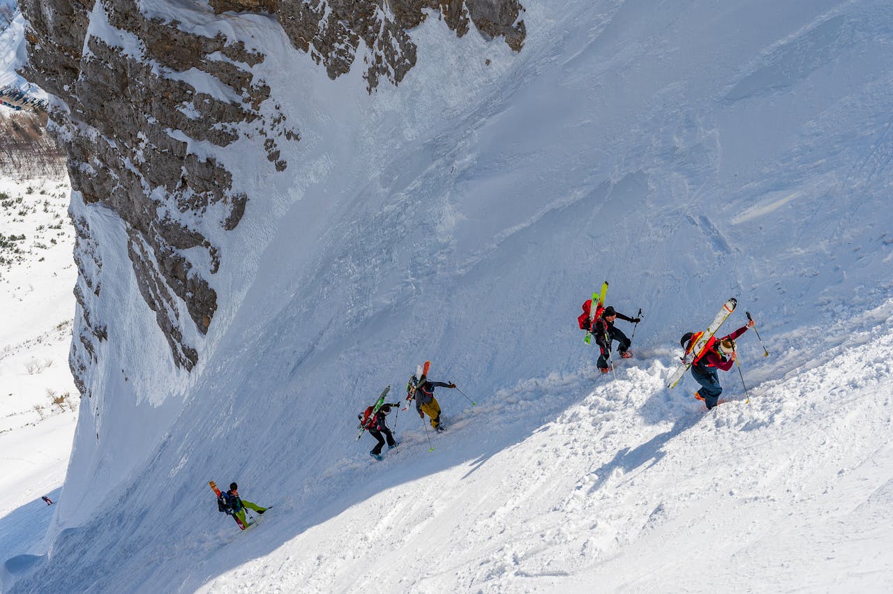 Adventurers hike up a steep, snowy mountain slope, carrying skis on a winter day.